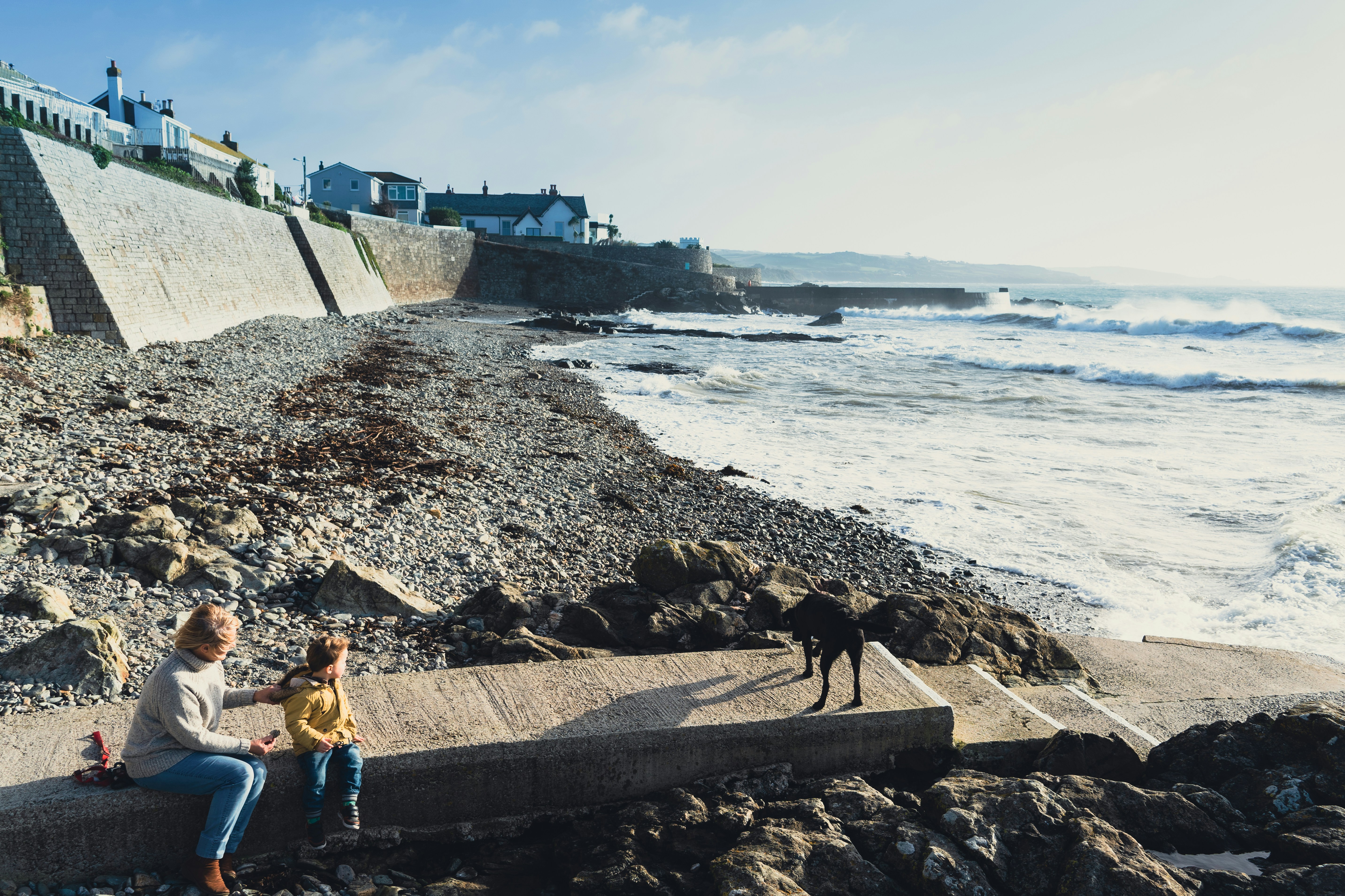 People sitting on the sea wall along the Penzance seafront with waves breaking on the rocky beach