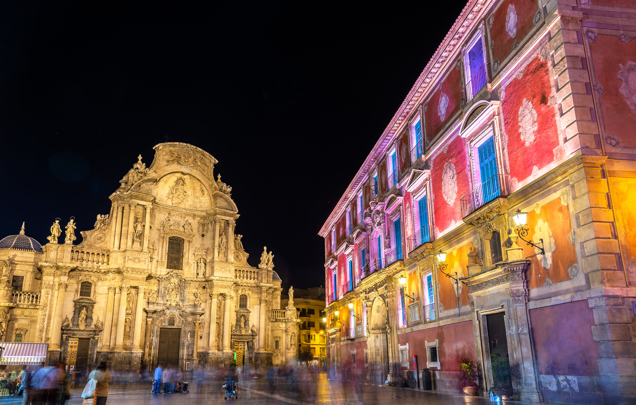 Murcia Cathedral and Palacio Episcopal illuminated at night, with people moving through Plaza del Cardenal Belluga.