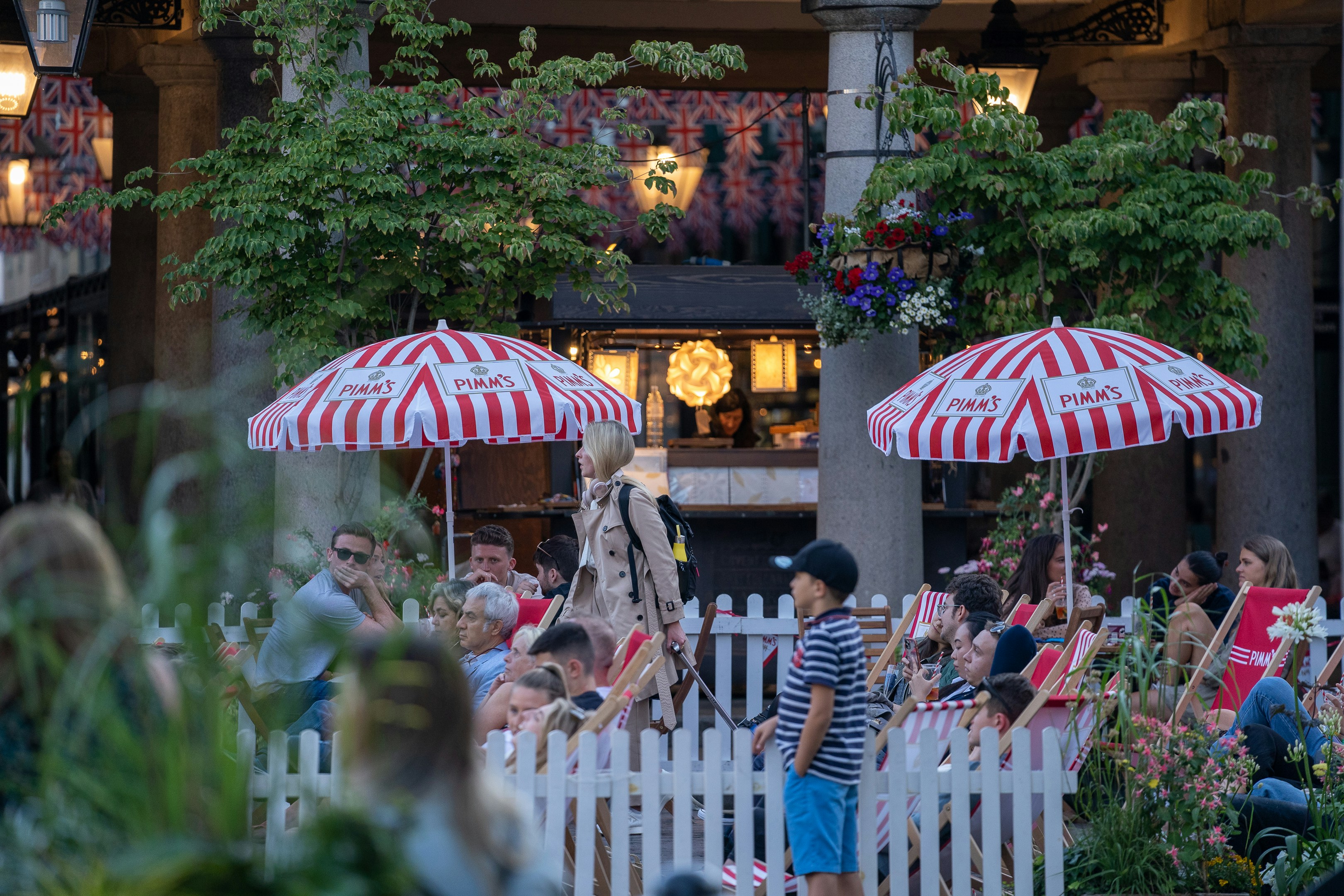 People sitting outside at a London pub terrace with red-and-white Pimm&rsquo;s umbrellas and deckchairs in summer.