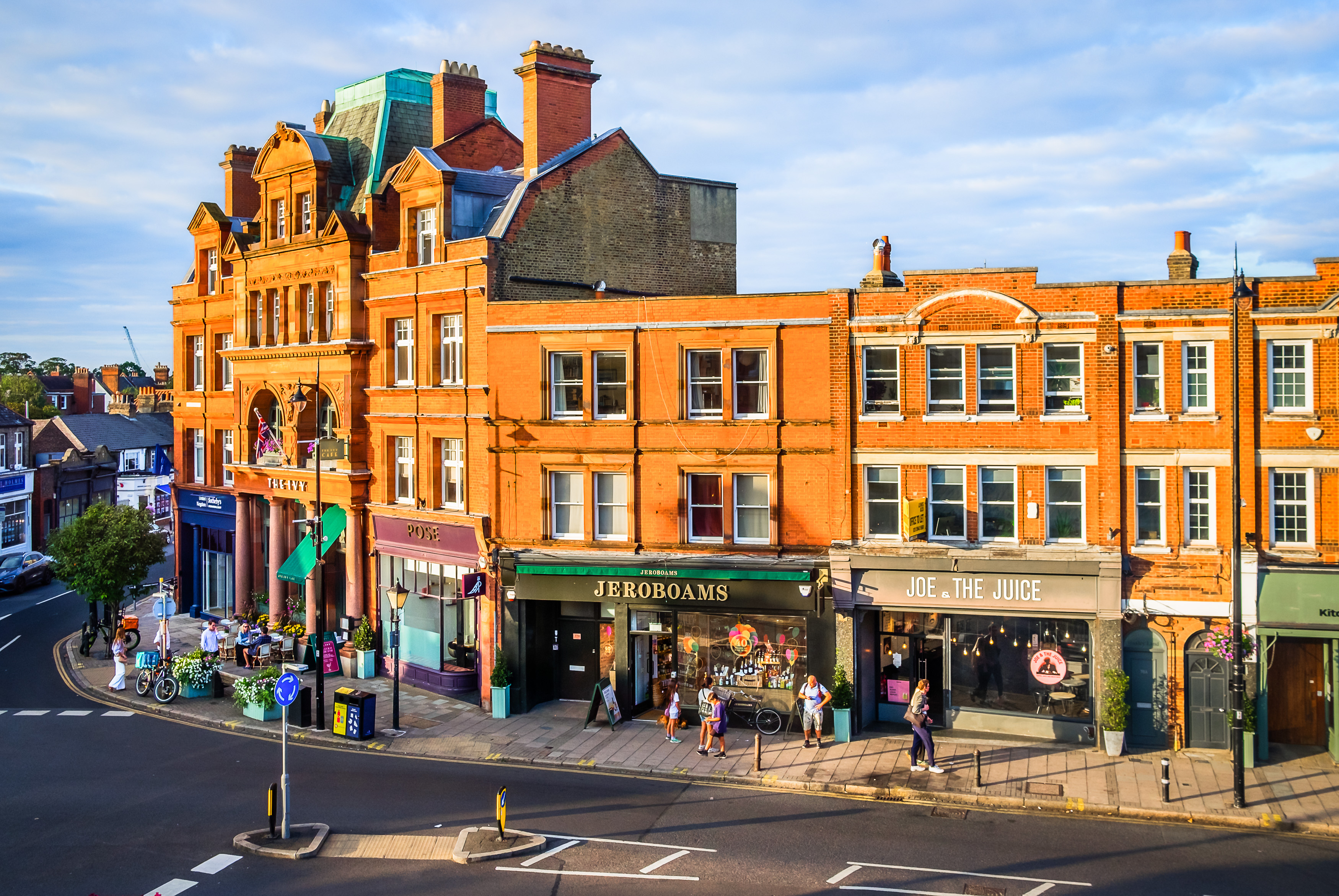 Wimbledon Village street with red-brick buildings, shops, caf&eacute;s and hotel frontage in warm evening light.